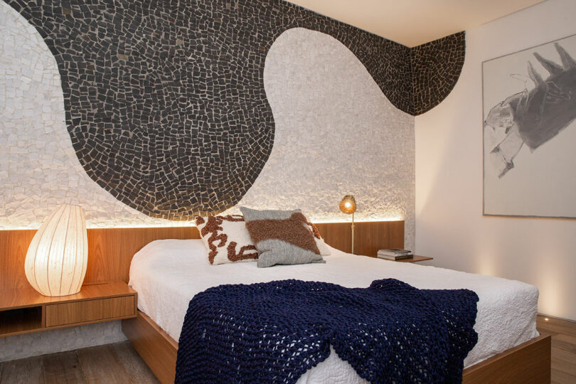 Modern bedroom with a double bed, white bedding, and a navy throw. The back wall features a black and white mosaic design. A wooden side table holds a spherical lamp. Artwork hangs on the right wall.