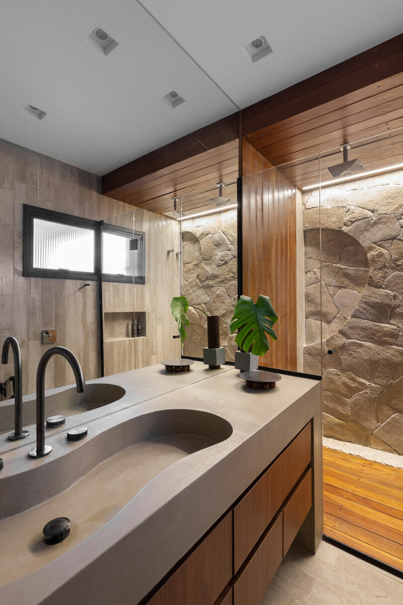 Modern bathroom with a double sink vanity, large mirror, and a shower area featuring stone walls. Two plants are placed on the vanity.