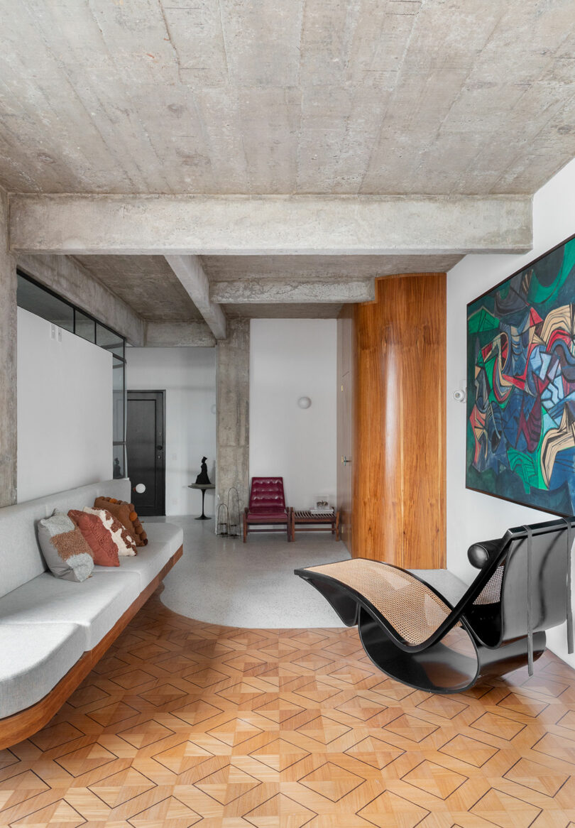 Modern living room with concrete ceiling, parquet flooring, a curved sofa with cushions, artistic wall painting, unique black chair, and decorative items including a small sculpture.