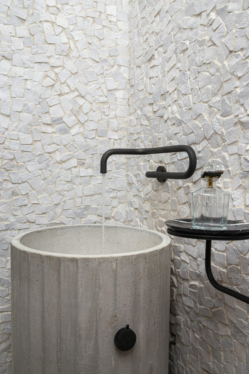 Concrete sink with a wall-mounted black faucet, running water, and a small table holding a glass and bottle, set against a textured white mosaic wall.