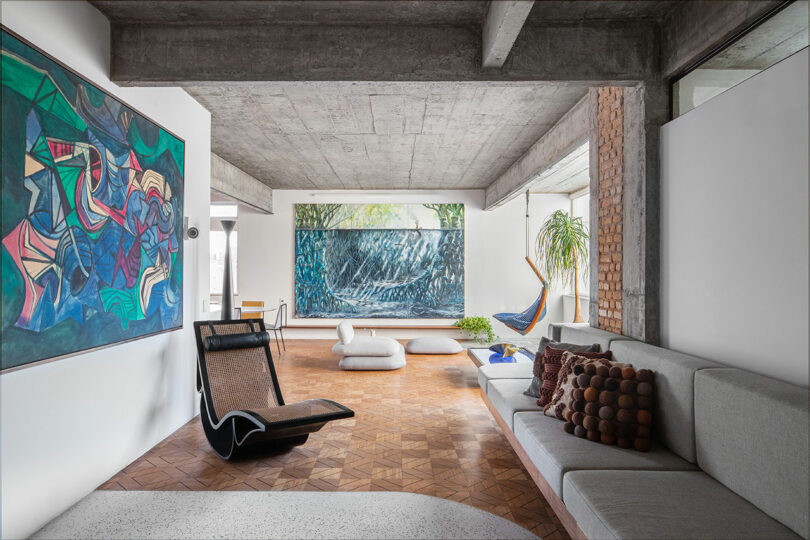 A modern living room with concrete ceiling, abstract artwork, a gray couch, geometric wall art, and wooden flooring. Large windows and plants enhance the natural light.