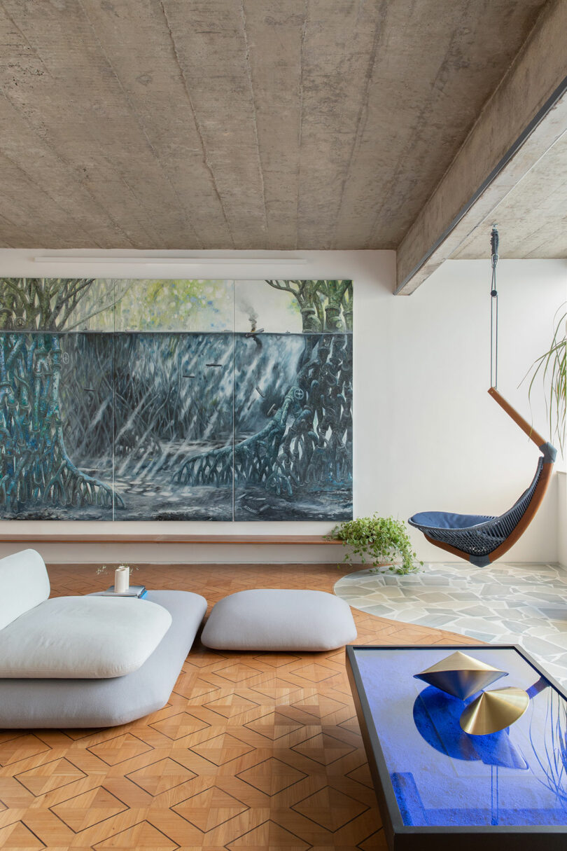 Modern living room with a large abstract mural, low seating, a glass coffee table, and a hanging chair. The floor is a mix of wood and tile, with a potted plant nearby.