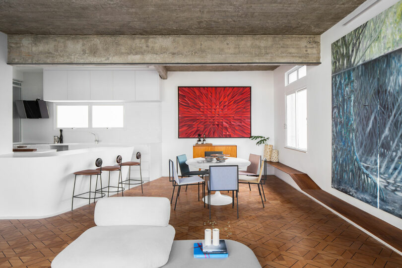 Modern open-concept living space with a white sofa, wooden flooring, dining set, abstract red artwork on the wall, and kitchen with barstools.