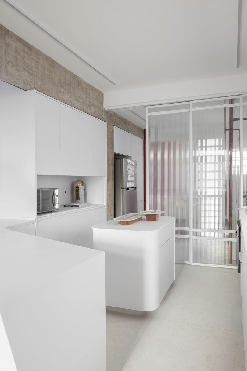 Modern kitchen with white cabinets, a center island, microwave, refrigerator, and a textured glass sliding door. Concrete ceiling and minimalistic design.