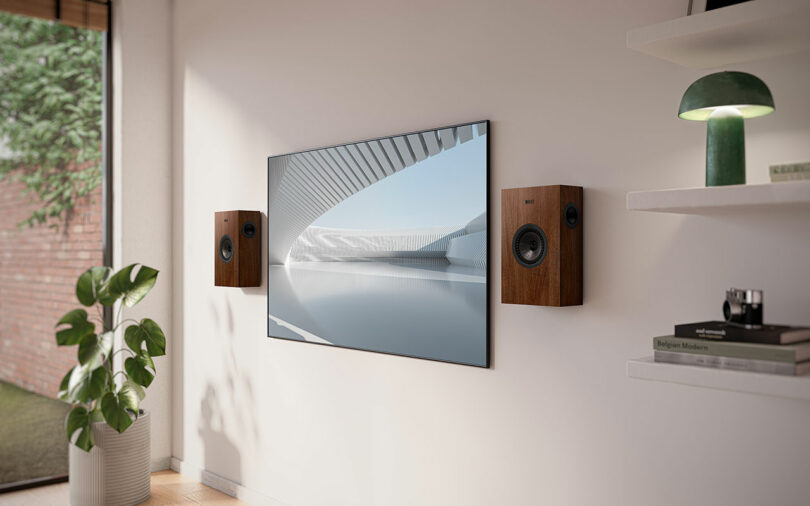 A wall-mounted flat-screen TV is flanked by sleek KEF speakers in a modern living room, featuring a potted plant and a green lamp elegantly placed on white shelves.