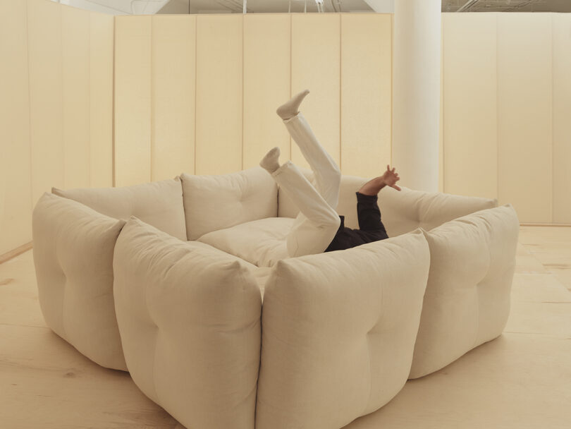 A person in white pants playfully reclines with legs up on a large, soft beige sofa in a minimalistic room.