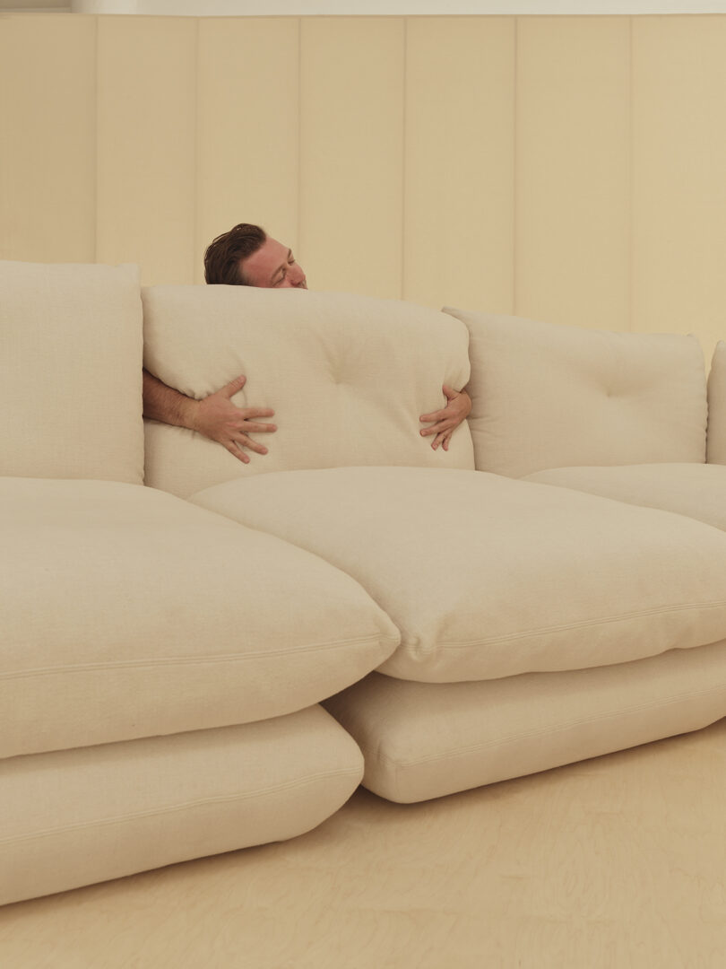 Person peeking over the back of a beige sofa, with hands gripping the large cushions.