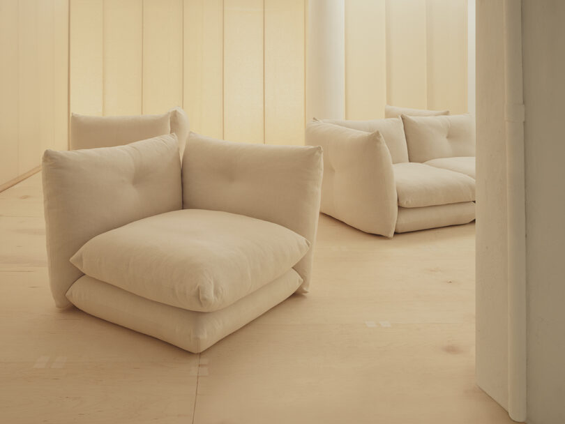 Two beige, cushioned lounge chairs and a matching loveseat in a minimalist room with light wooden flooring and soft lighting.