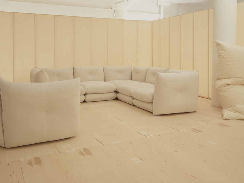 A beige modular sofa sits in a corner of a room with light-colored wood flooring and beige walls. A large, soft cushion is on the right.