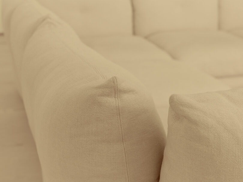 Close-up of a beige fabric sectional sofa, showing the texture and stitching on the cushions and armrest.