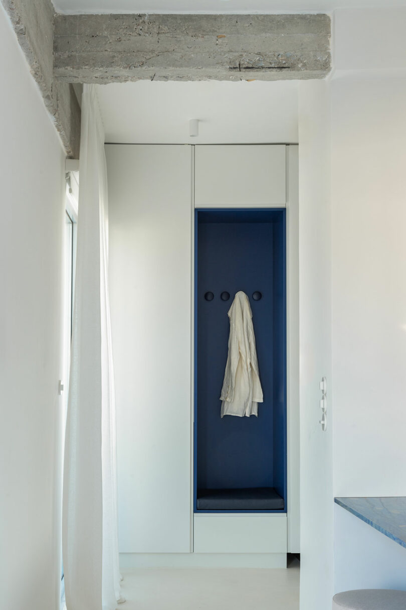 A minimalist alcove with a white coat hanging on hooks, surrounded by light-colored walls and ceiling, featuring a raw concrete beam above.