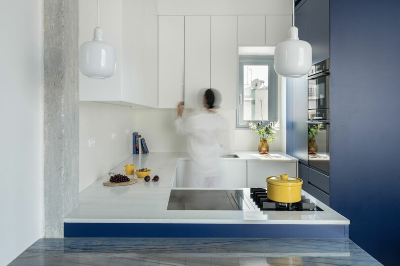 A person in motion stands in a modern kitchen with white cabinets, blue accents, and a yellow pot on the stove.