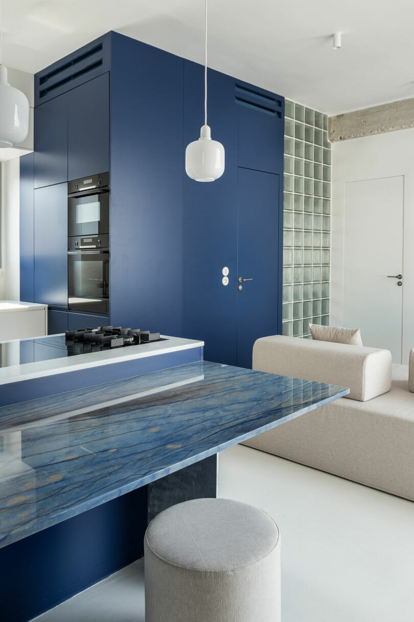 Modern kitchen and living area with a blue and white color scheme, featuring a blue marble dining table, blue cabinetry, built-in appliances, and a beige sofa with a glass block wall.