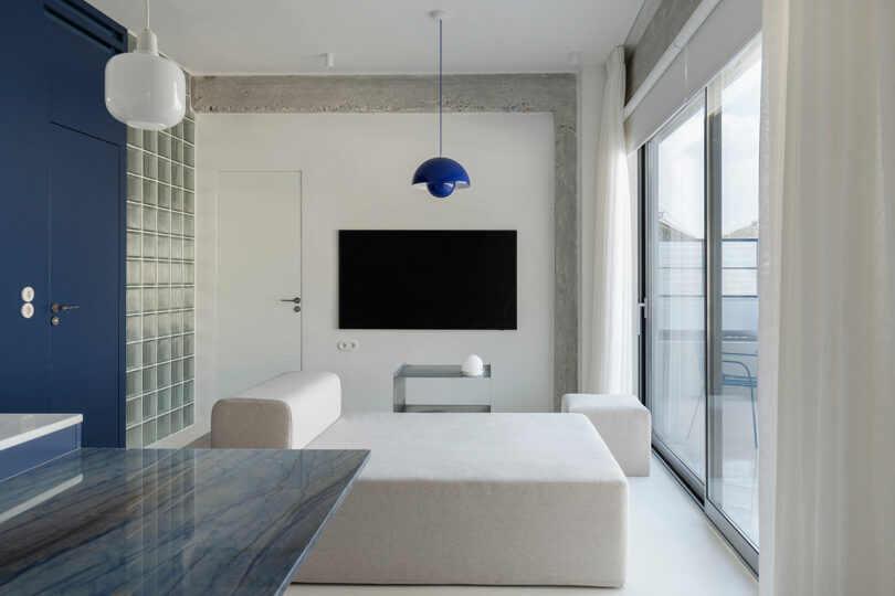 Modern living room with a minimalist design features a beige sofa, flat-screen TV, blue pendant light, glass block wall, and large sliding glass doors with sheer curtains.