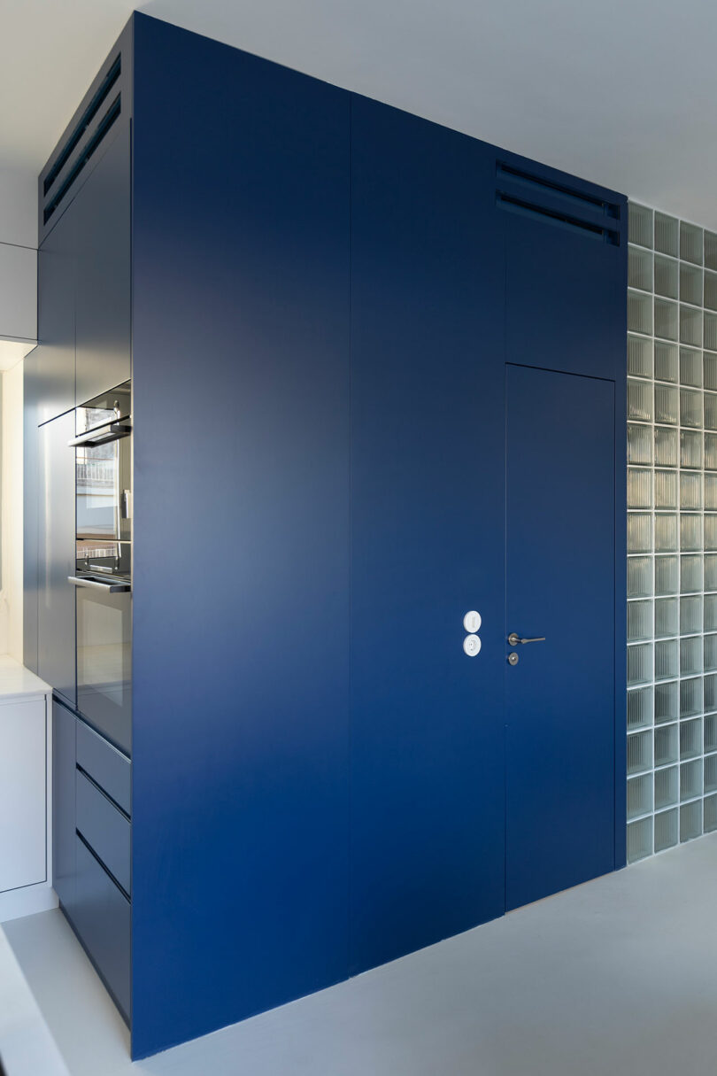 Solid blue kitchen cabinet with integrated oven and drawers, adjacent to a wall of glass blocks.