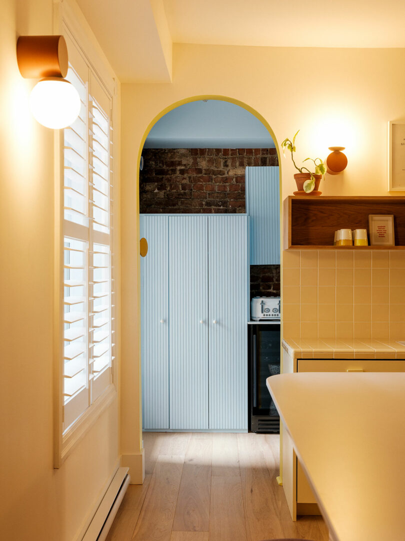 A cozy kitchen with yellow walls, a white countertop, and warm lighting. A wooden floor leads to an archway revealing light blue cabinetry and a brick accent wall. A small plant is on a shelf.