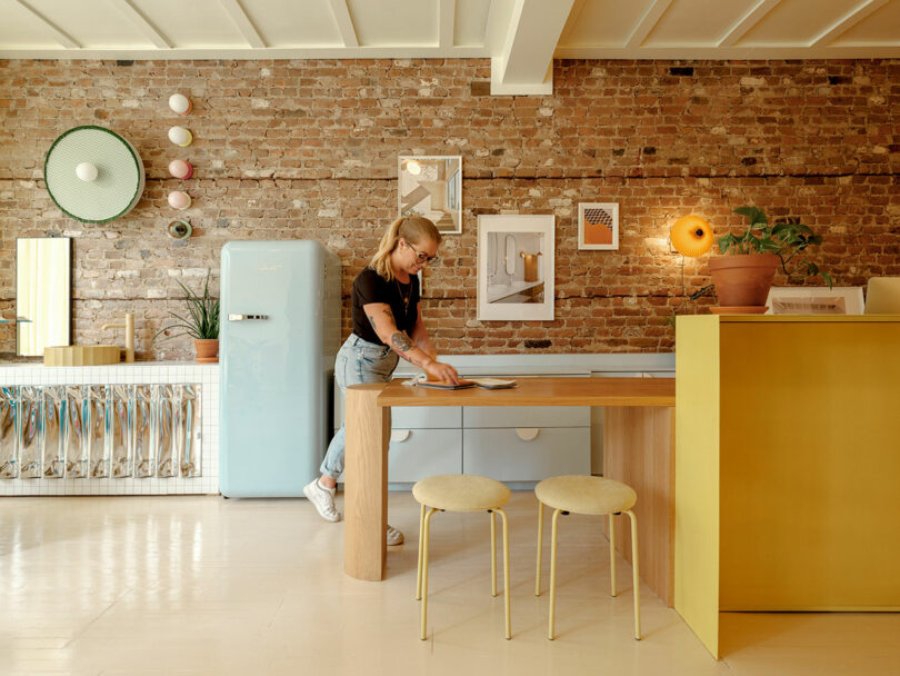 A woman stands in a bright kitchen with a brick wall, blue fridge, and wooden counter. Decor includes artwork and mirrors, with two beige stools in the foreground.