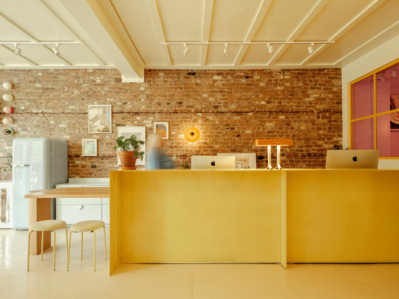 Modern office space with a long yellow desk, two iMacs, a potted plant, and a blurred person walking. Exposed brick wall with framed art, beige stools, and a retro fridge in the background.