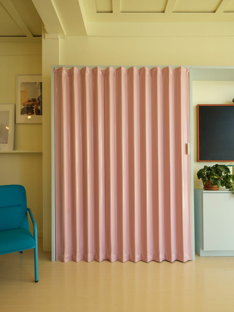 A room with a pink folding partition, a teal chair on the left, framed pictures on the wall, and a plant on a cabinet under a blackboard.