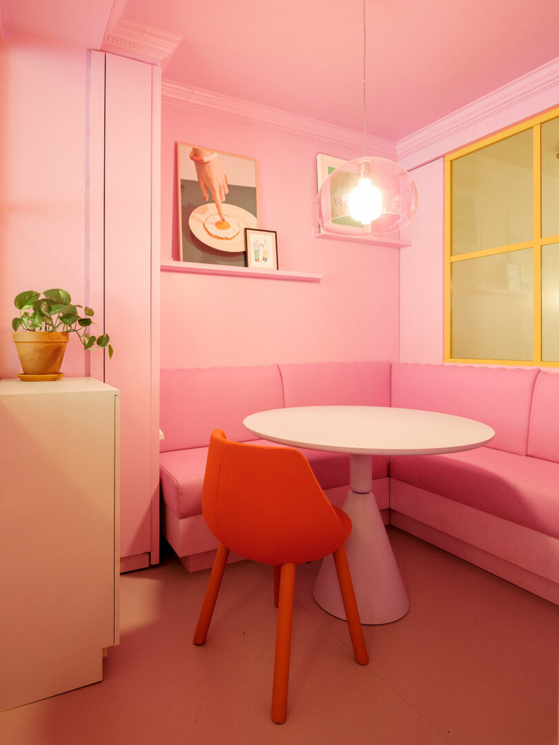 A small pink dining nook with a round white table, red chair, and corner banquette seating. A hanging pendant light and framed pictures adorn the space. A plant sits on a white cabinet.