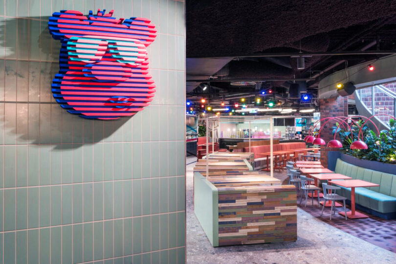 Colorful restaurant interior with eclectic decor, featuring a geometric bear sculpture on a tiled wall, vibrant seating, and multicolored lighting.