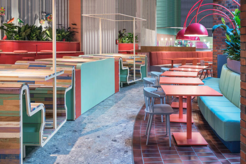 Colorful modern restaurant interior with wooden tables, cushioned seating in various pastel shades, red accents, and lush greenery.