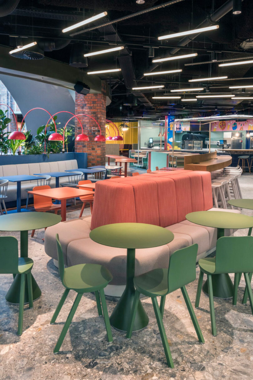 Modern, colorful cafe interior with green tables, red and white chairs, benches, and overhead lights. Plants and decorative lamps add to the vibrant atmosphere.