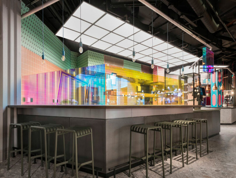 A modern bar with metal stools, geometric ceiling lights, and colorful glass walls.