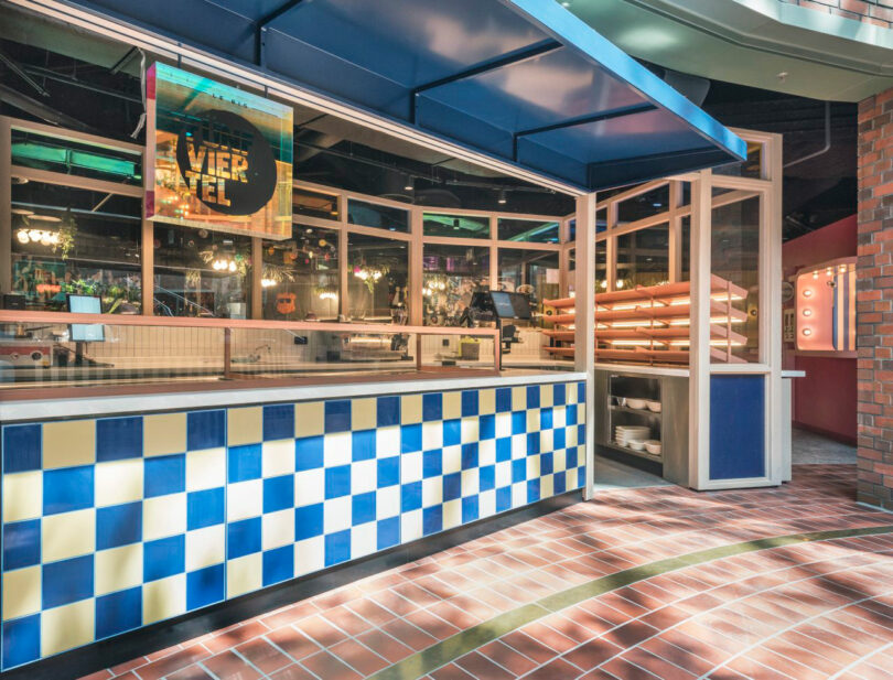 Restaurant interior with a counter featuring a blue and yellow checkered pattern, open shelving, and a menu sign above. Lights and tables are visible in the background.