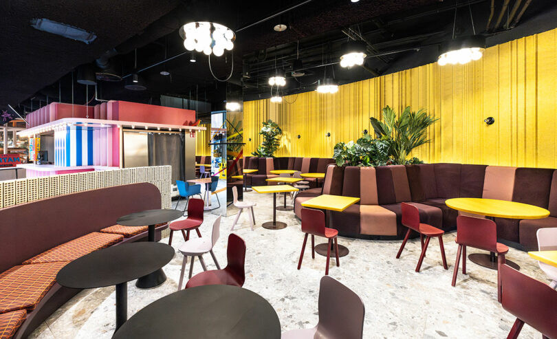 A modern cafe interior with colorful chairs and round tables, yellow wall panels, and plants. Bright lighting and a contemporary design.