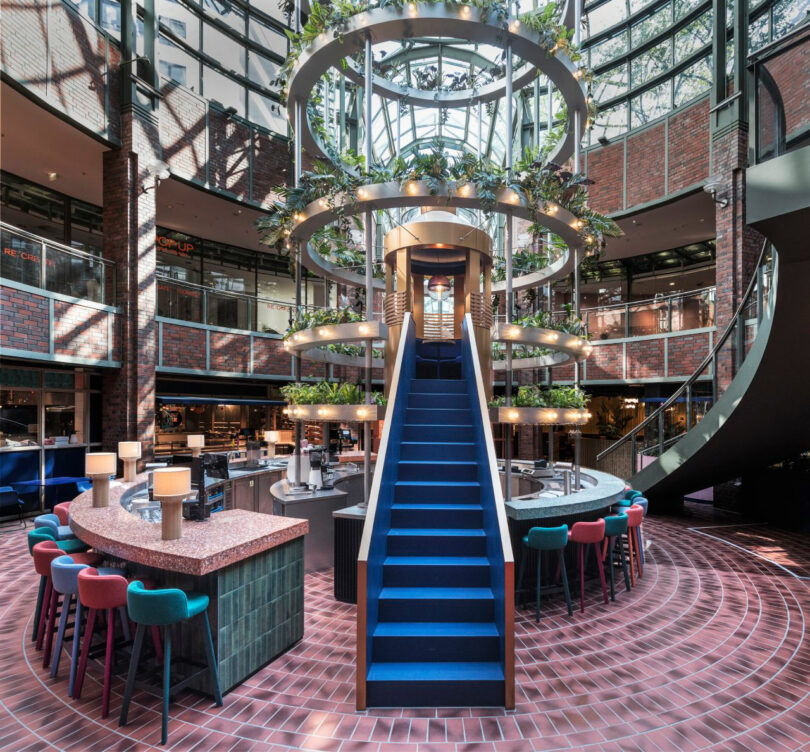 A modern, circular restaurant interior with a central staircase, surrounded by colorful seating and hanging greenery under a glass ceiling.