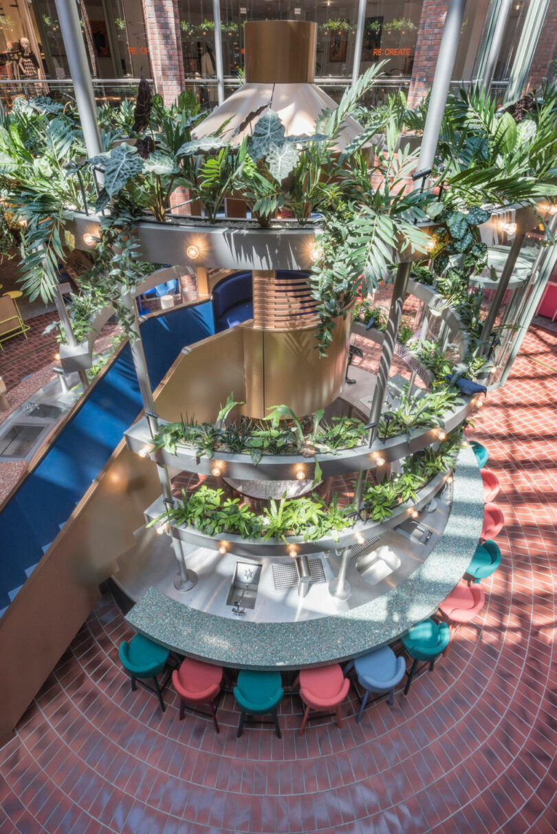 Overhead view of a circular bar with surrounding greenery, colorful bar stools, and a vibrant, modern architectural design in a spacious indoor setting with natural light.