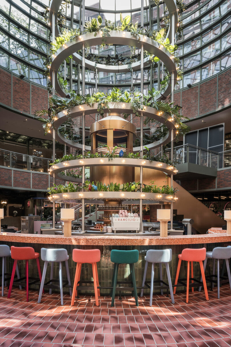 Circular bar with colorful stools and tiered plant displays under a glass dome ceiling.