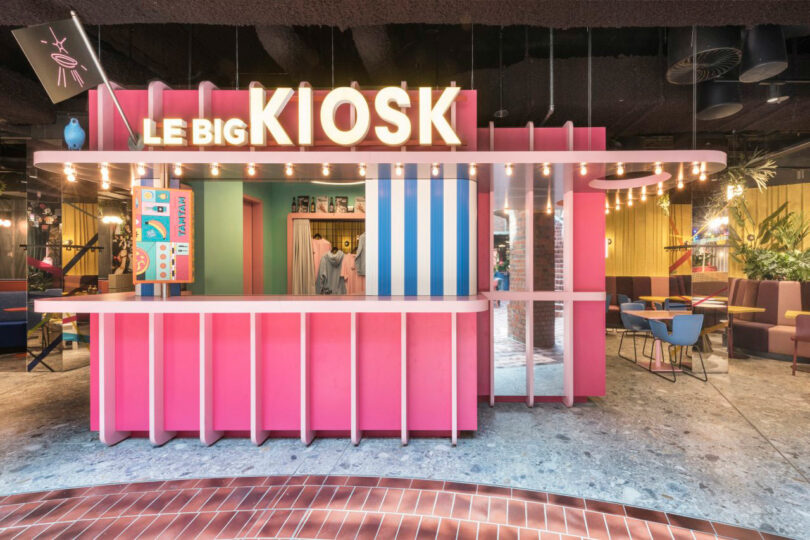 Colorful kiosk with a prominent "LE BIG KIOSK" sign, featuring a pink and blue design, surrounded by seating areas and illuminated by numerous hanging lights.