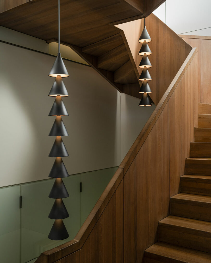 Wooden staircase with glass railing, featuring modern, black cone-shaped pendant lights hanging from the ceiling, designed in the elegant style of Legato by Luca Nichetto.