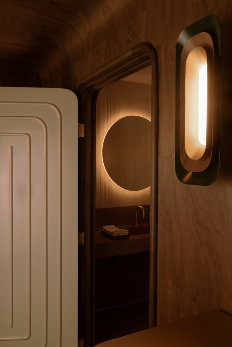 Warmly lit bathroom with wood-paneled walls. Circular mirror above a sink reflects soft light. Door partially open, showing a modern wall light nearby.