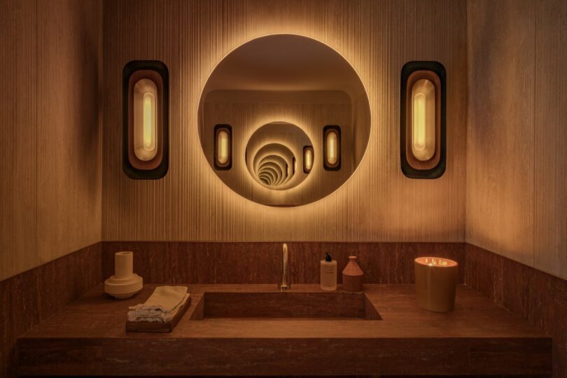 A modern bathroom with a circular mirror creating an optical illusion. Two wall lights flank the mirror. The countertop has a sink, folded towels, soap dispenser, and a candle.