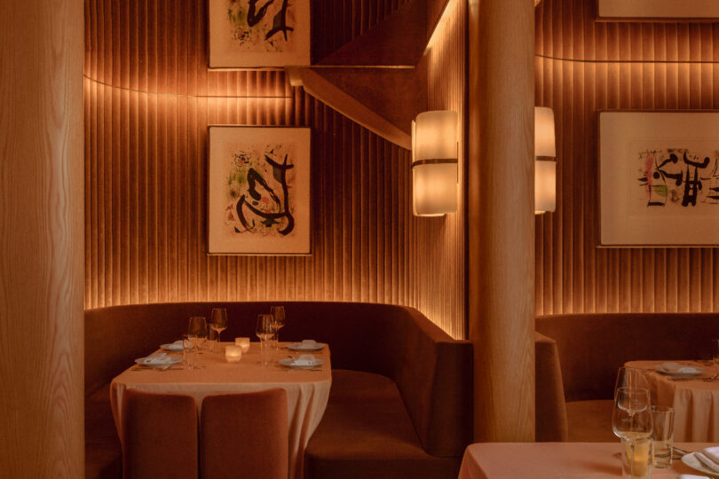 Cozy restaurant interior with warm lighting, round booth seating, framed abstract art on walls, and tables set with white tablecloths and glassware.