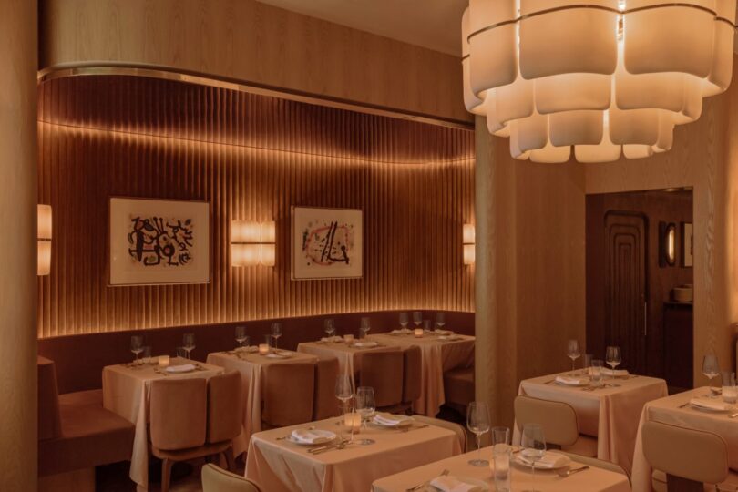 A warmly lit restaurant interior with tables set for dining. Wooden walls feature framed abstract art, and a large geometric chandelier hangs from the ceiling.