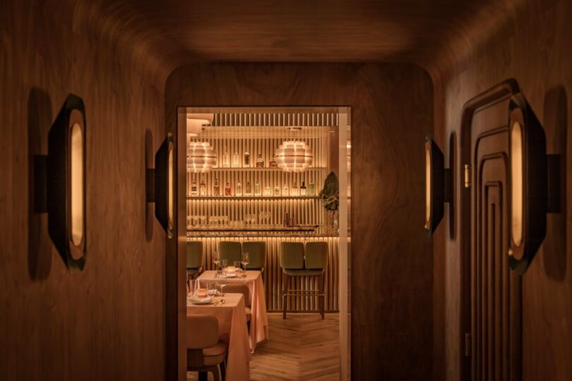 Dimly lit restaurant interior with a warm, wooden hallway leading to a dining area with tables, chairs, and a bar. Soft lighting from wall sconces and overhead fixtures creates an inviting atmosphere.