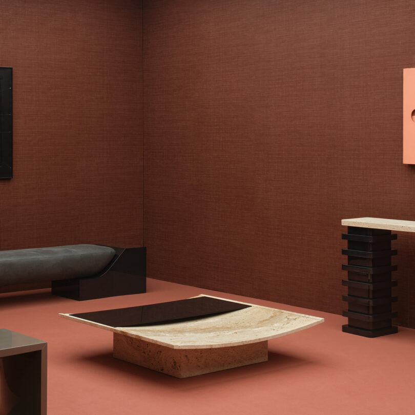 A minimalist room with brown walls and a beige coffee table embodies the Marks of Existence Movimento, featuring a black cushioned bench and an artistic console table with layered black supports.