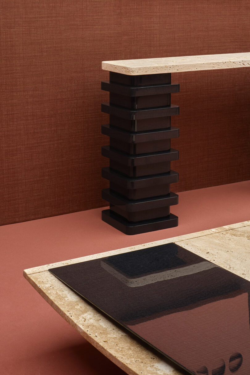 Modern table with a polished stone top and a black stacked pedestal base, set against a brown textured wall and red floor.