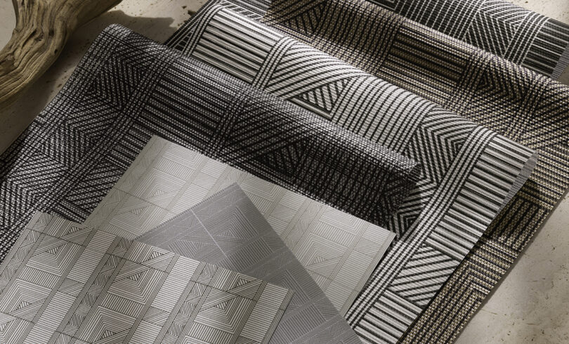 Four textured, geometric-patterned fabrics in varying shades of black, gray, and beige are laid out overlapping on a surface. A piece of driftwood is partially visible in the corner.