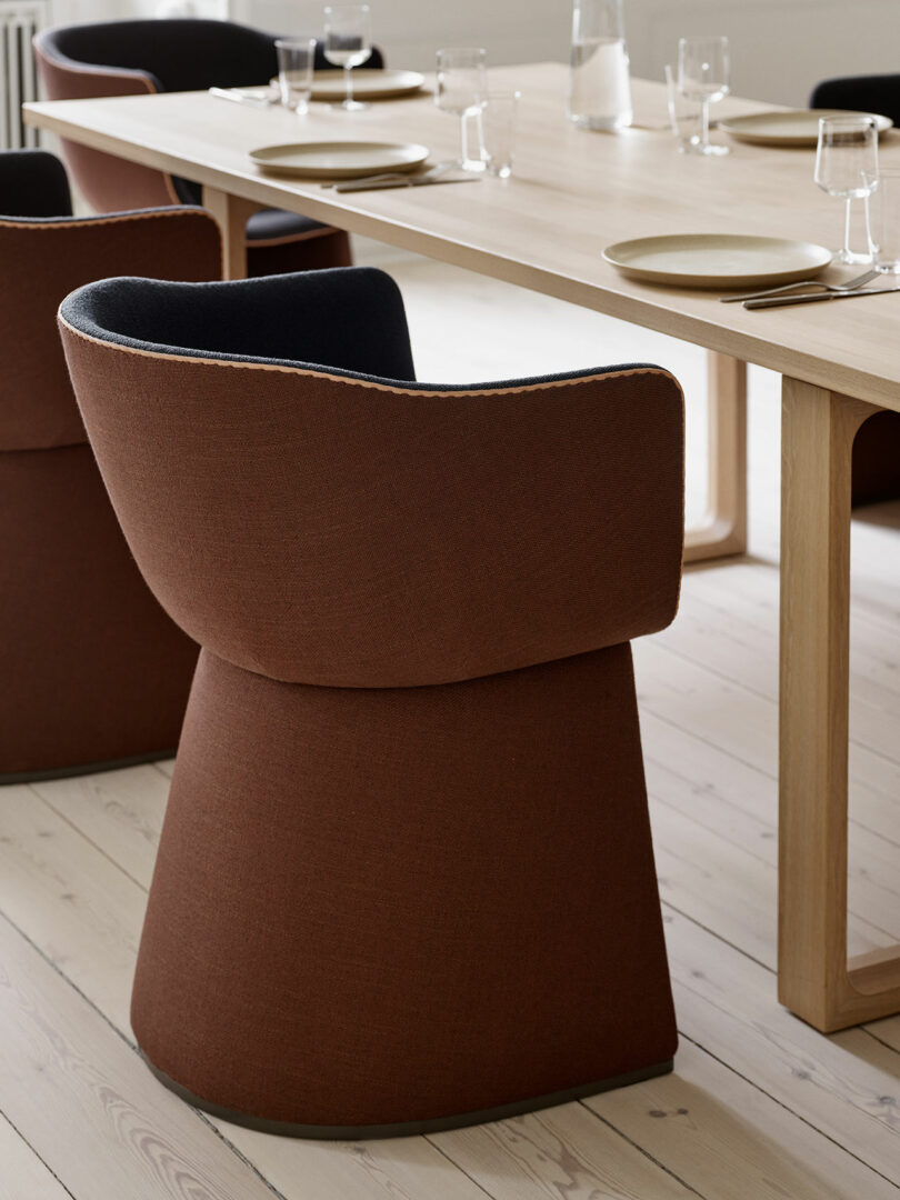 A modern dining room with a light wood table, set with glassware and plates, surrounded by brown upholstered chairs on a light wood floor.