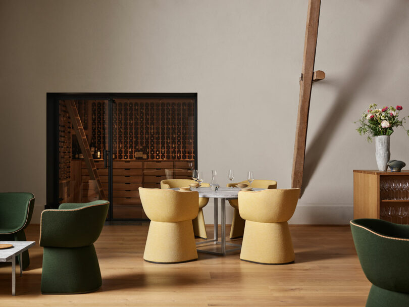 A modern, minimalist dining area with yellow and green chairs around a table, a wine rack in the background, and a vase of flowers on a sideboard.