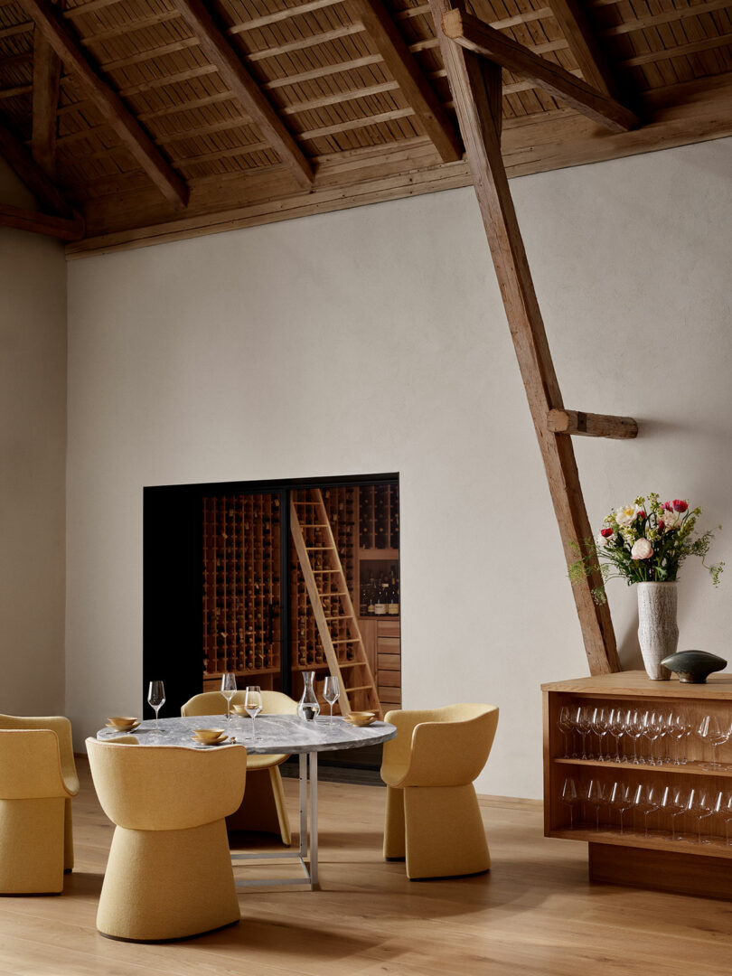 A dining room with a round table set for four. Yellow chairs surround the table. A wooden wall with wine storage and a ladder is visible. A cabinet with flowers and glasses is on the right.