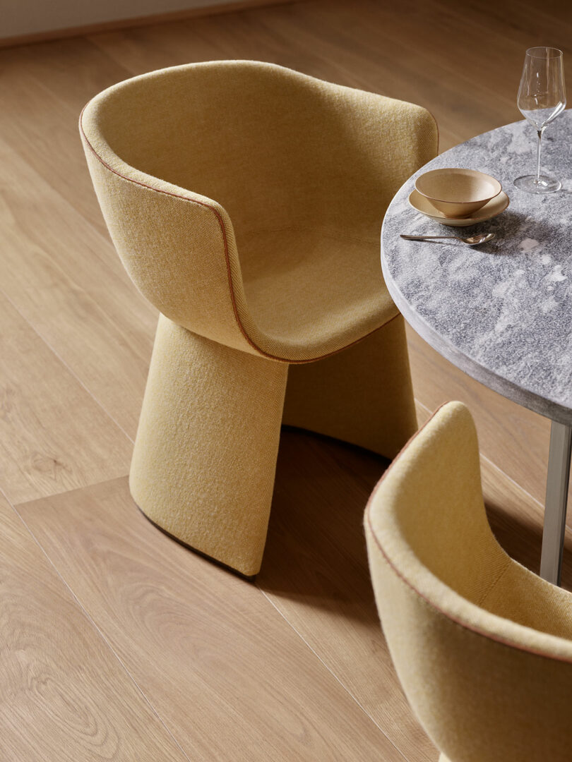 Two modern yellow chairs are positioned around a round marble table. A wine glass and bowl with a spoon are set on the table, and the floor is wooden.