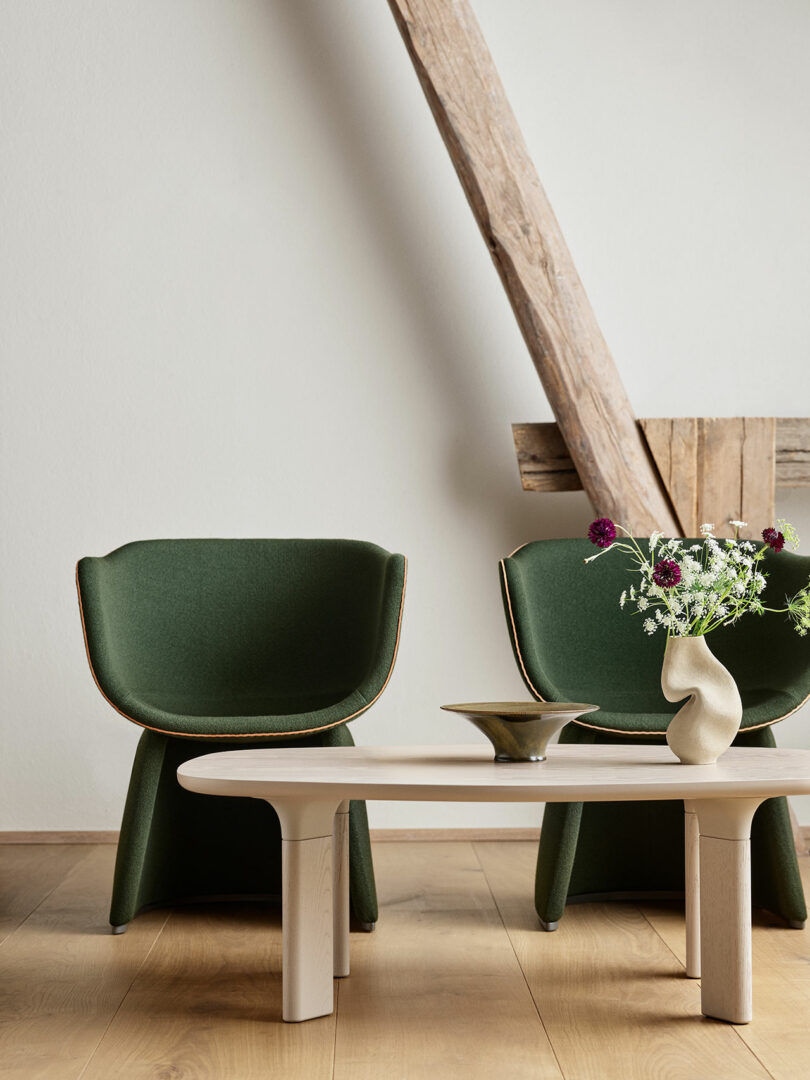 A minimalist room with two green chairs, a light wooden table, and a simple floral arrangement in a vase. The setting features wood accents and a light, neutral color scheme.