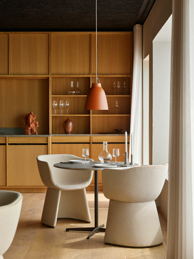 A modern dining area with two cream chairs, a round table set with glasses, a carafe, and candles, under a hanging orange lamp. Wooden shelves with decor fill the background.