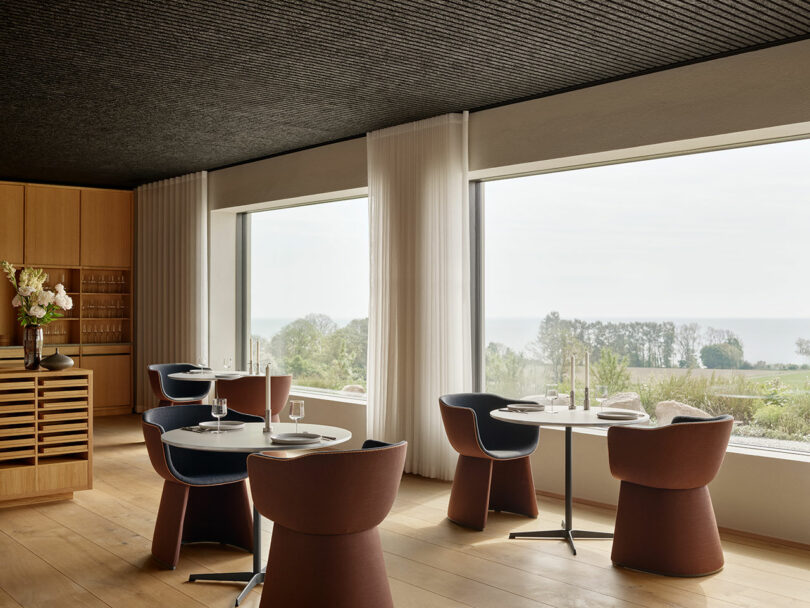 Modern dining room with round tables, brown chairs, and large windows overlooking a scenic outdoor view. Wood paneling and light curtains enhance the minimalist decor.